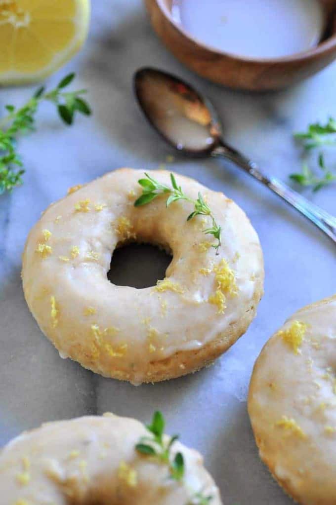 three vegan lemon donuts on a marble counter top with fresh rosemary, a lemon, a spoon, and a small bowl of glaze on the side 