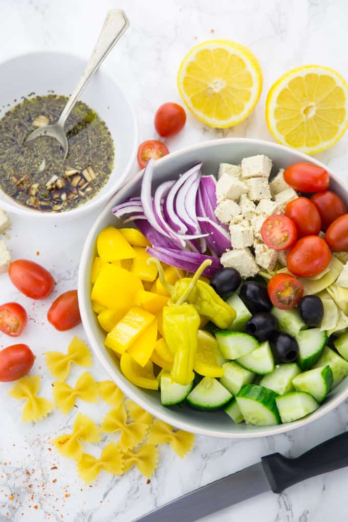 ingredients for a Greek pasta salad with cut-up lemons, cherry tomatoes, farfalle pasta, and Greek vinaigrette in a small bowl on the side