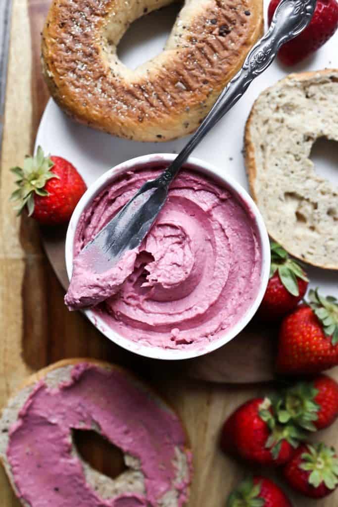 a bowl of vegan strawberry cream cheese with a knife on top and two bagels and fresh strawberries on the side 