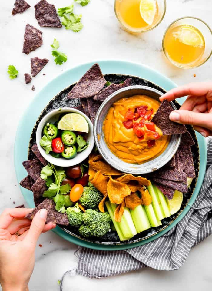 a blue plate with nachos, celery sticks, broccoli and a bowl of vegan queso with someone dipping a nacho into it 