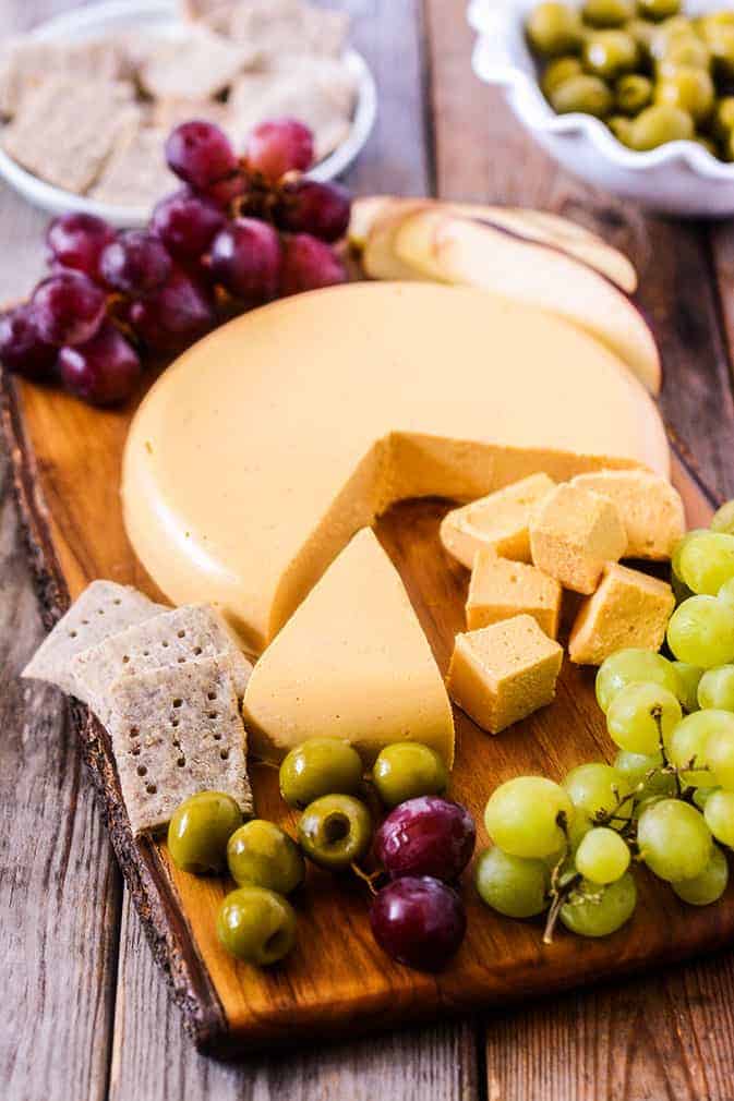 Vegan cheddar cheese on a wooden board with crackers, grapes, and olives on the side 