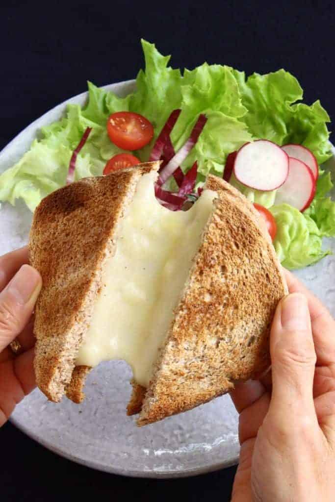 someone holding two halves of a vegan cheese sandwich over a plate with a salad with melted cheese between both halves 