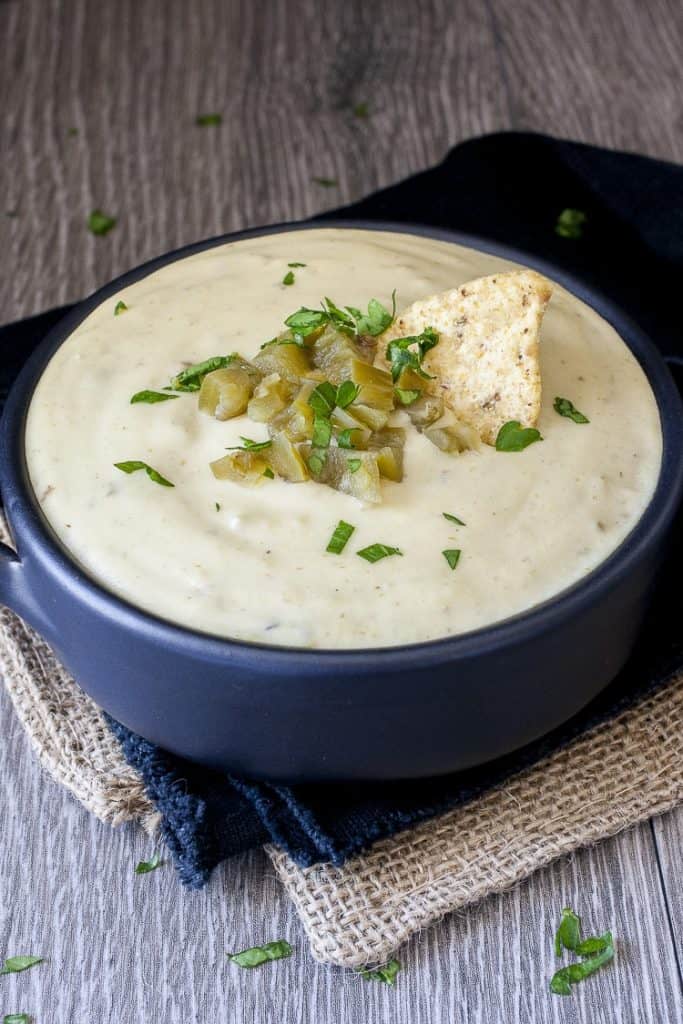 vegan queso blanco in a blue bowl on a wooden counter top with green chilis and a nacho chip on top 