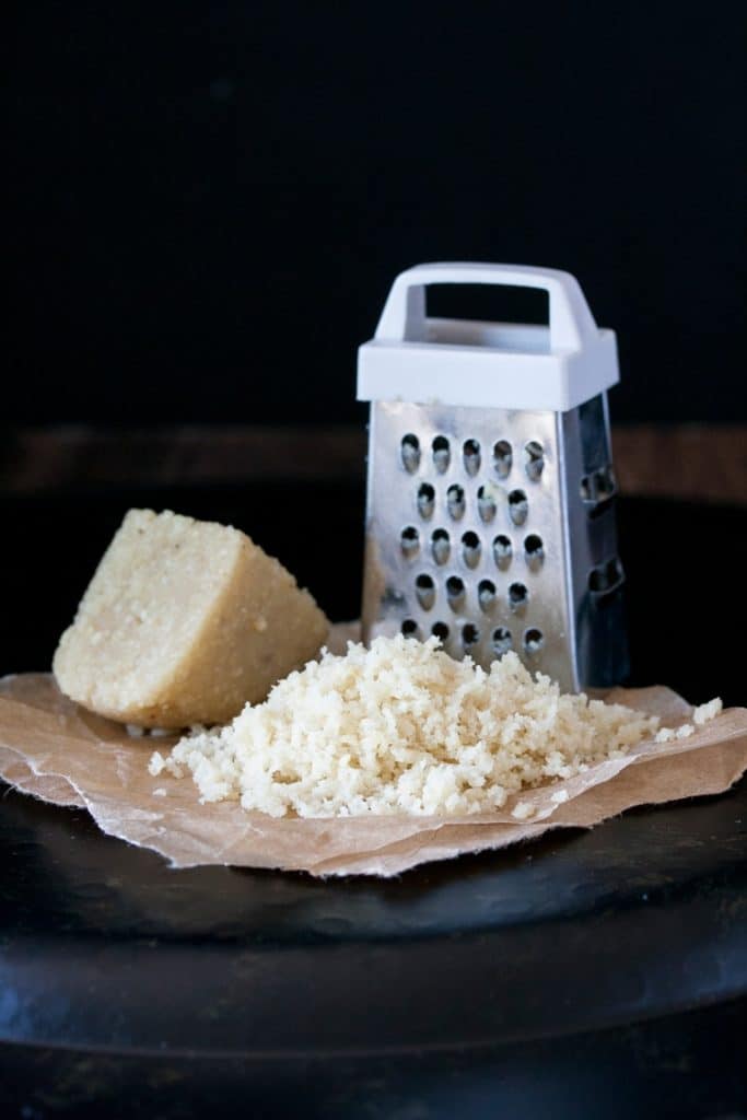 a piece of vegan Parmesan cheese with a tiny cheese grater on a black surface with a black background 