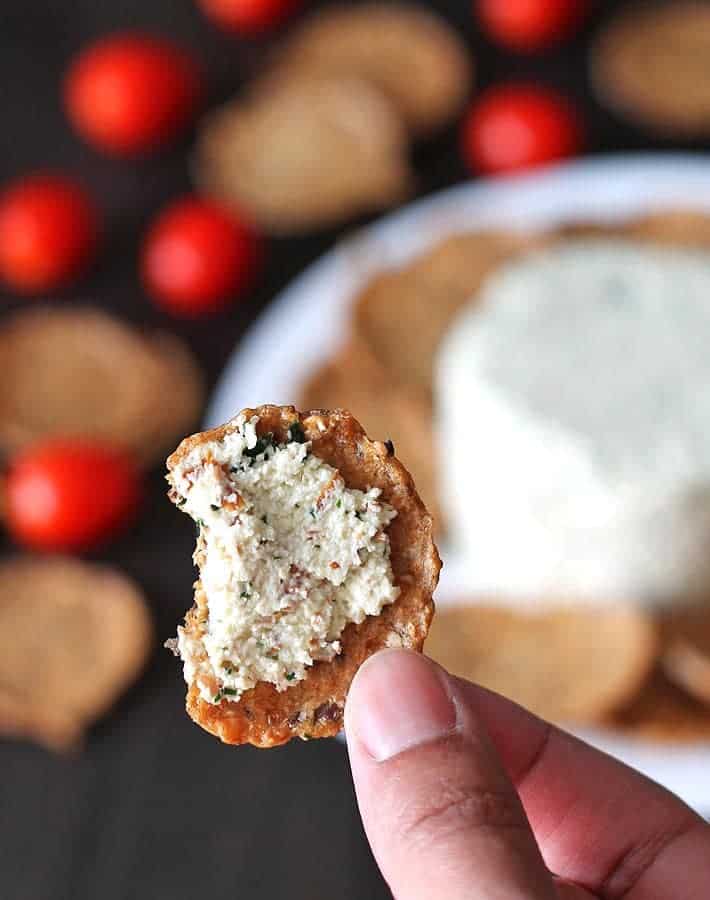 a hand holding a cracker with vegan cream cheese with cherry tomatoes and crackers in the background 