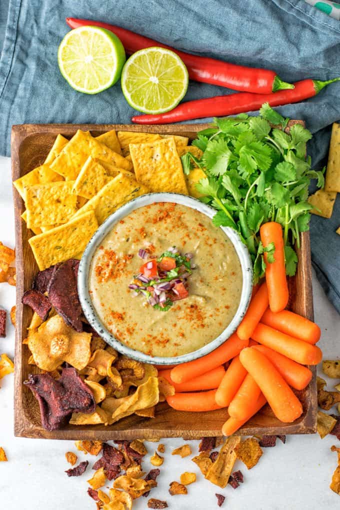 a bowl of vegan cheese dip on a wooden plate with crackers, baby carrots, vegetable chips, and cilantro on the side 