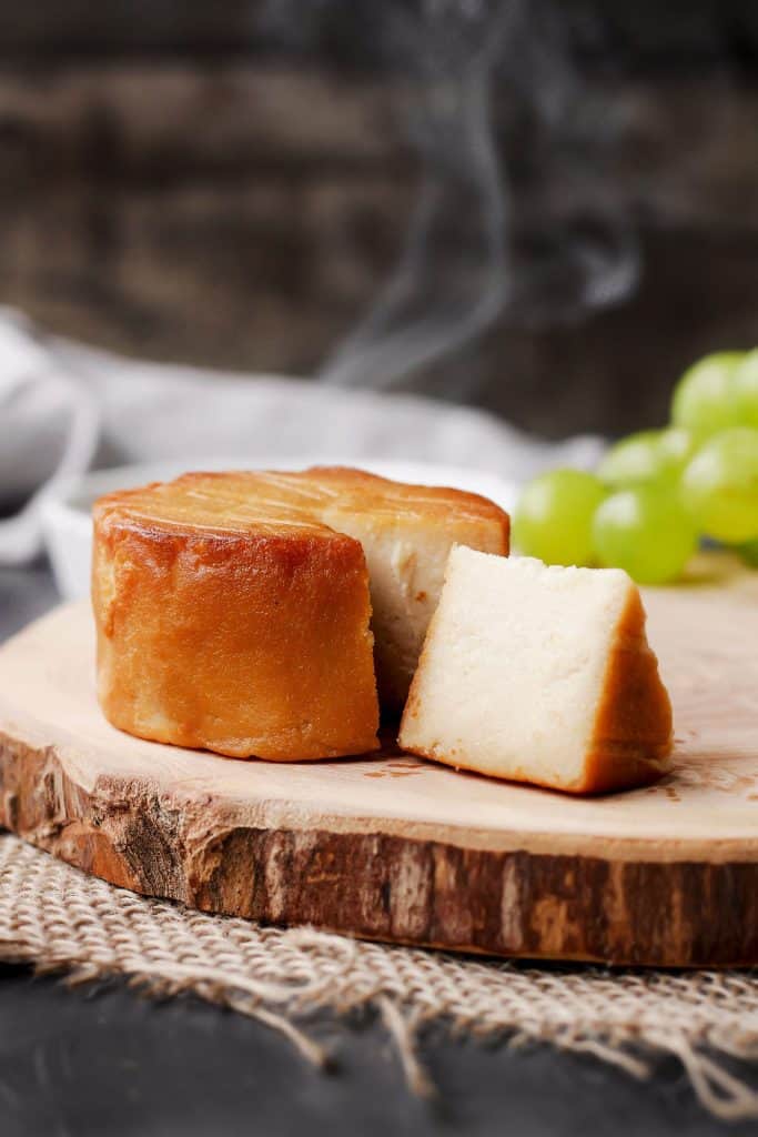 a piece of hickory smoked vegan cheese on a wooden board with grapes and smoke in the background 