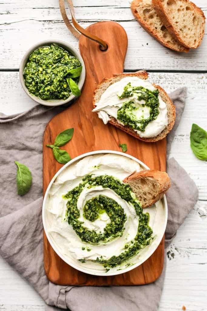 vegan pesto swirl cream cheese in a white bowl on a wooden board with slices of bread on the side and a small bowl of pesto on the side