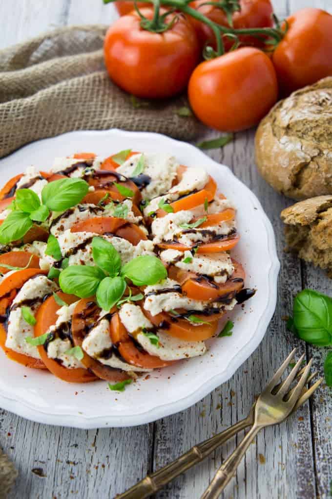 Vegan mozzarella cheese on a white plate with tomato slices and basil and whole tomatoes and bread in the background 