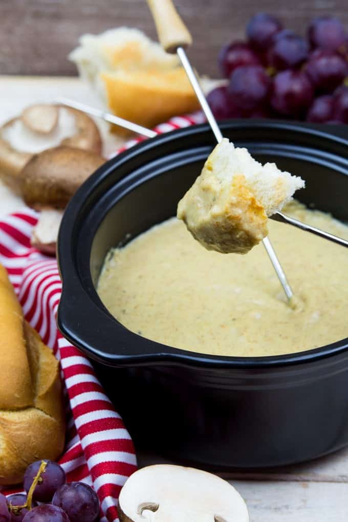 vegan cheese fondue in a black pot with someone dipping a piece of baguette into the cheese sauce and grapes, mushrooms, and more baguette in the background