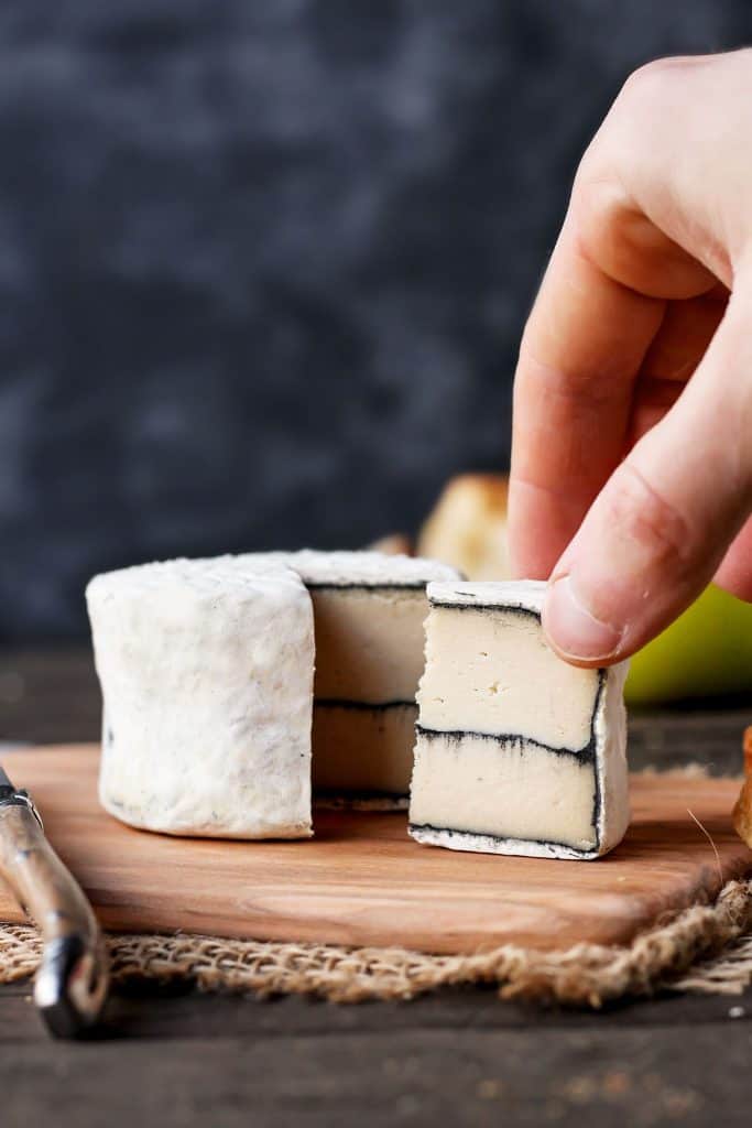 a piece of vegan ash ripened camembert on a wooden board with a knife on the side 