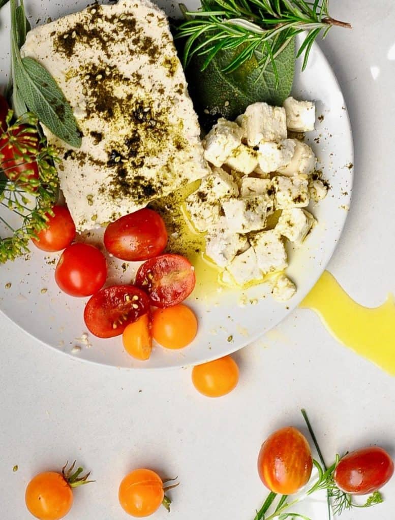 marinated almond feta on a white plate with cherry tomatoes, oil, and fresh herbs on the side 