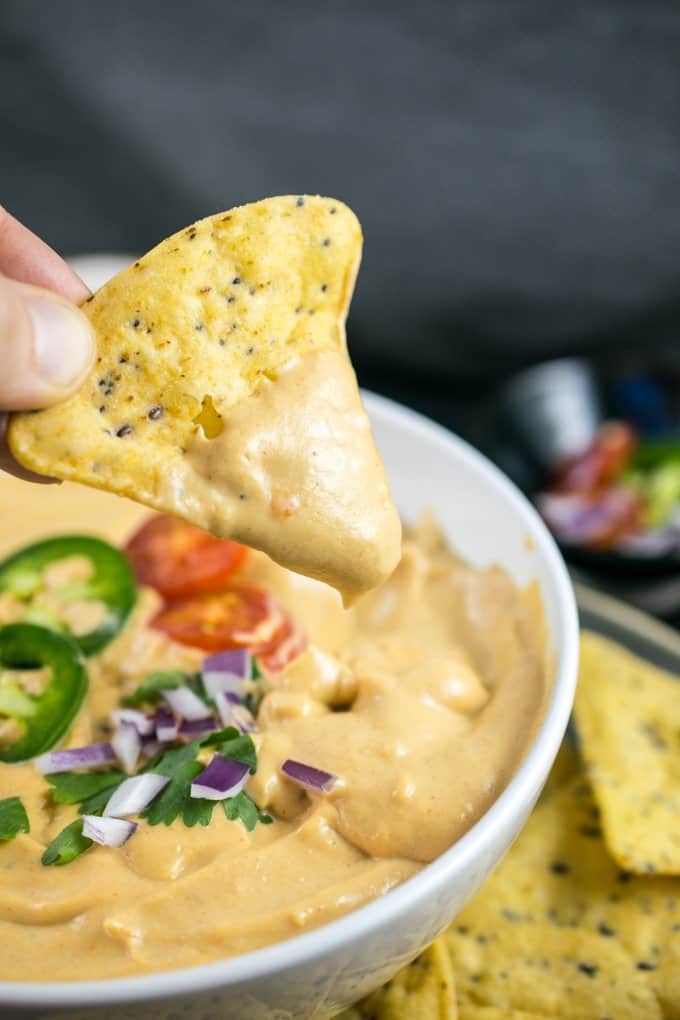 a hand dipping a nacho into a bowl of vegan queso with a black background 