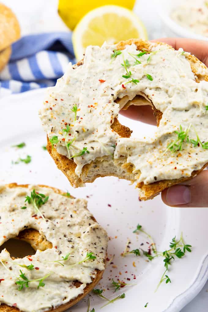 a hand holding a bagel with vegan cream cheese on a white plate