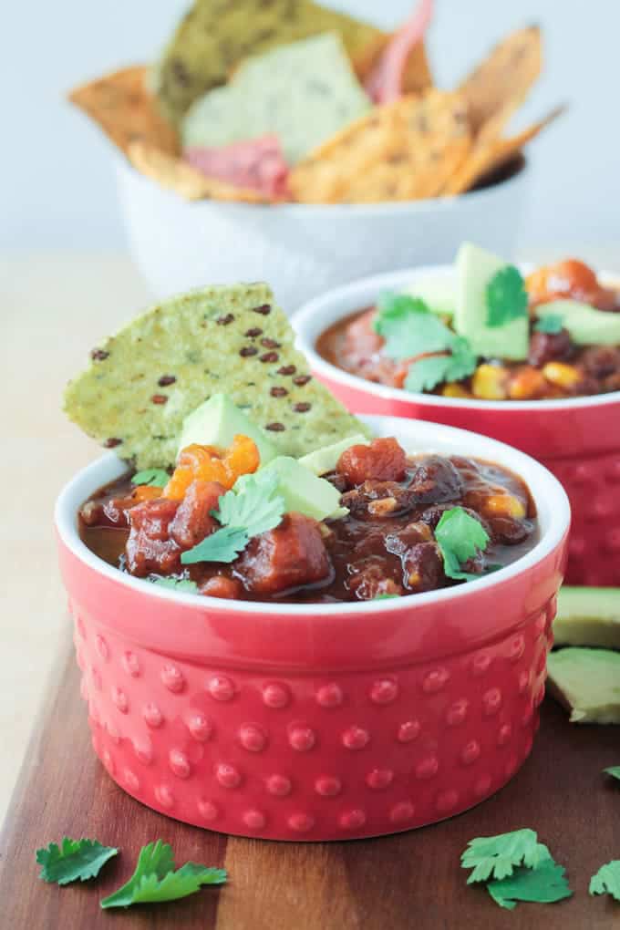 vegan chili with black beans and butternuts in two red bowls with a bowl of tortilla chips in the background