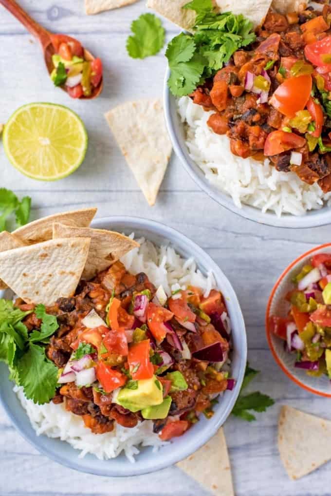 two bowls of vegan chili on a white surface with tortilla chips and a lime on the side