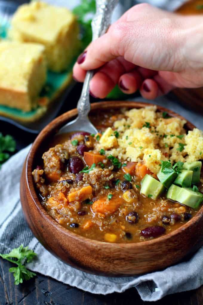 A hand with a fork in a wooden bowl of vegan quinoa chili with couscous on the side