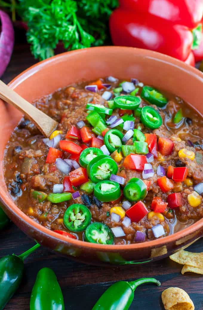 vegan chili with lentils in a brown bowl with a wooden spoon and a red bell pepper and fresh herbs in the background
