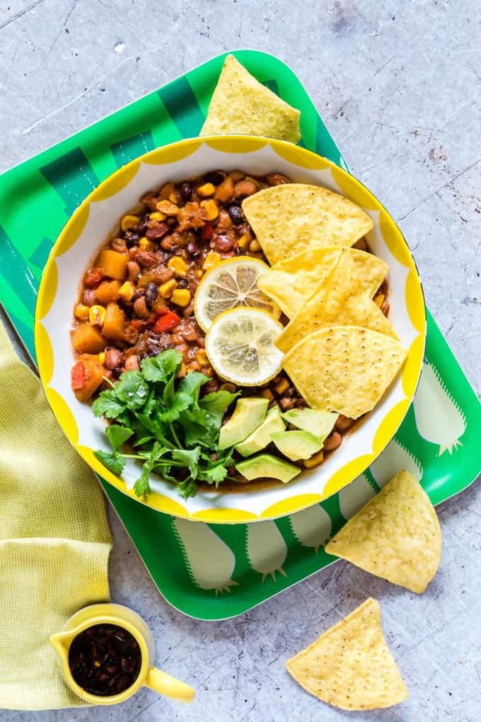 slow cooker vegan chili in a yellow bowl on a green tray with tortilla chips on the side