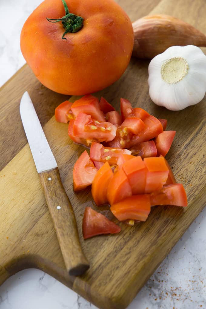 diced tomatoes, a knife, a clove of garlic. and a shallot on a wooden chopping board 
