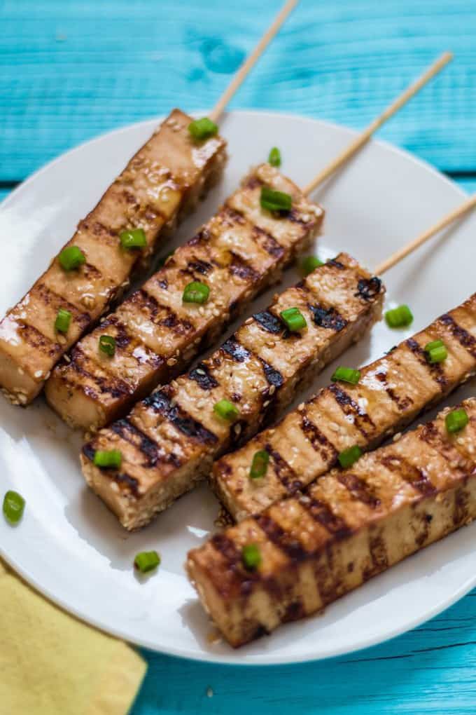 five tofu skewers on a white plate on a blue wooden surface with green onions sprinkled on top