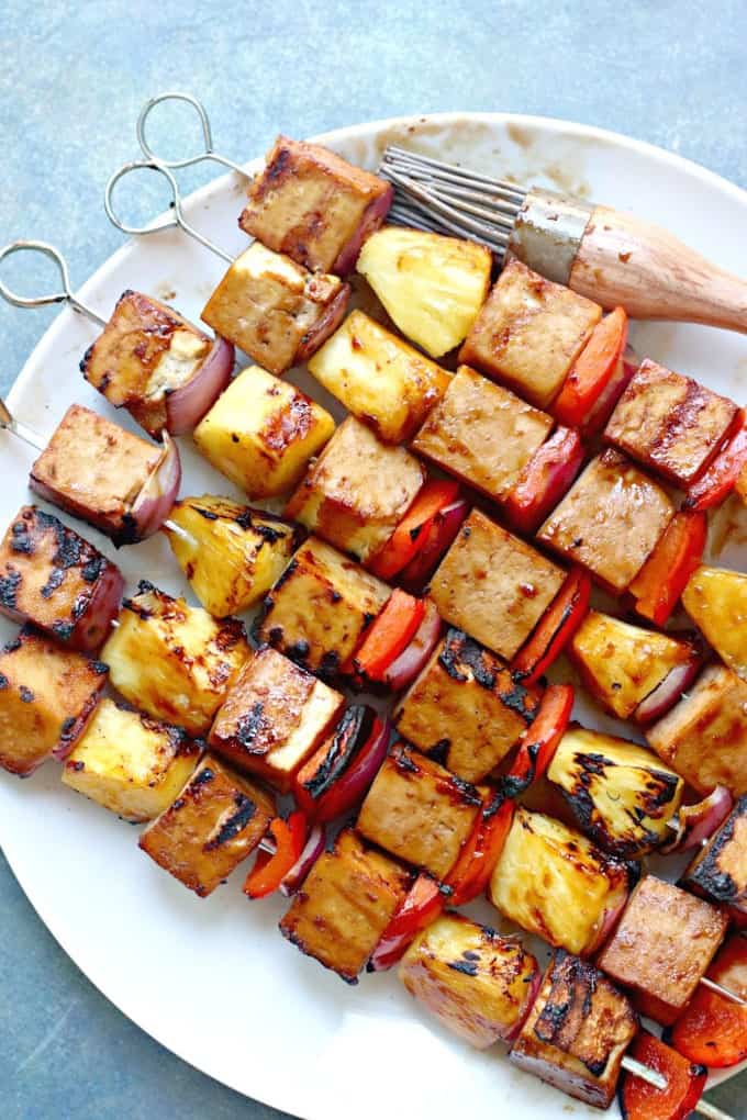 six tofu kabobs one a plate with a pastry brush on a blue countertop