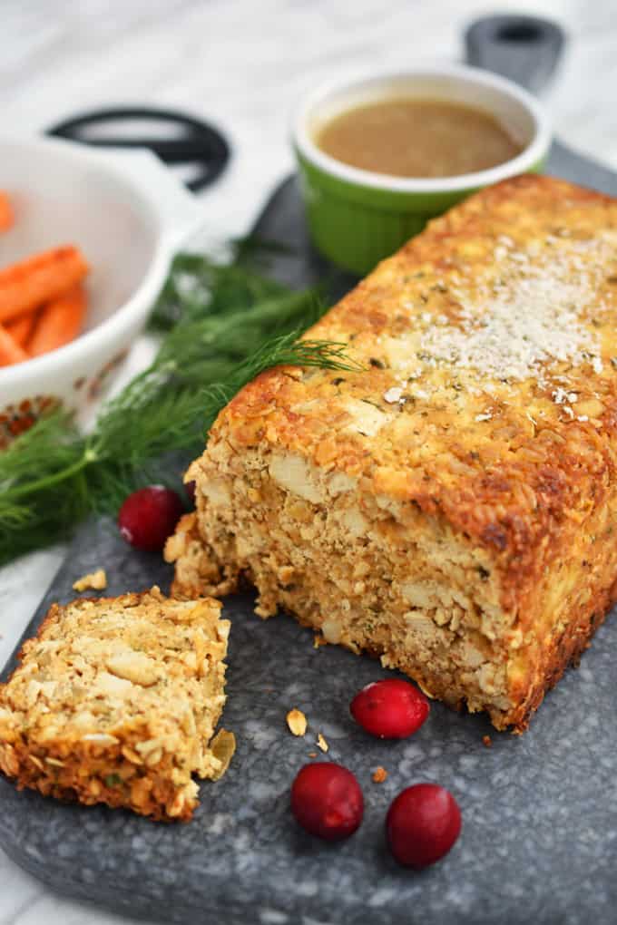 a tofu loaf on a dark serving board with a slice of the tofu loaf in the front and a bowl of sauce in the background