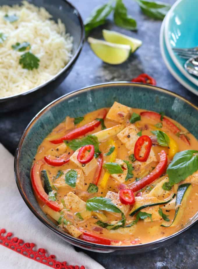 Thai curry with tofu in a blue bowl with a bowl of rice and lime wedges in the background