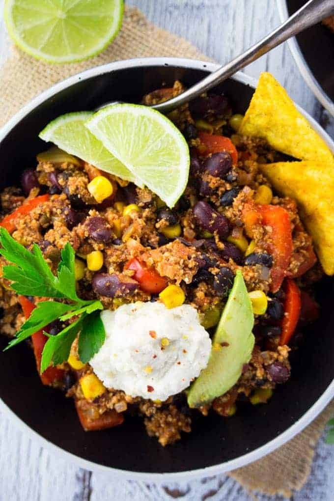 Tofu chili in a black bowl on a white wooden counter top with cashew sour cream and lime wedges on top