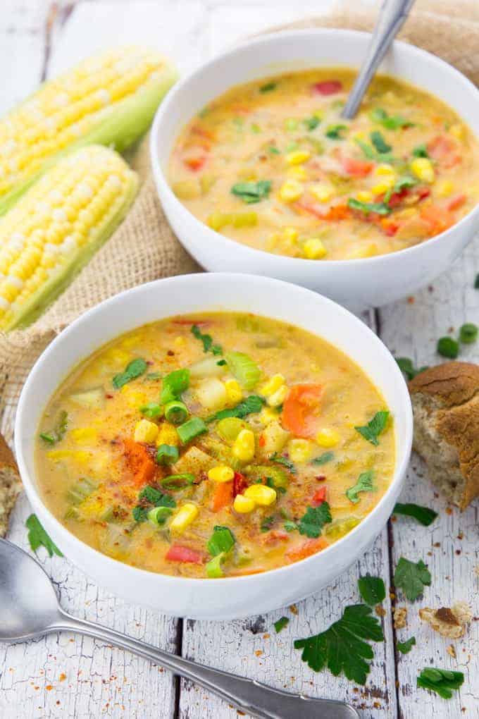 vegan corn chowder in two white bowls with spoons on a white wooden surface with corn cobs and bread in the background