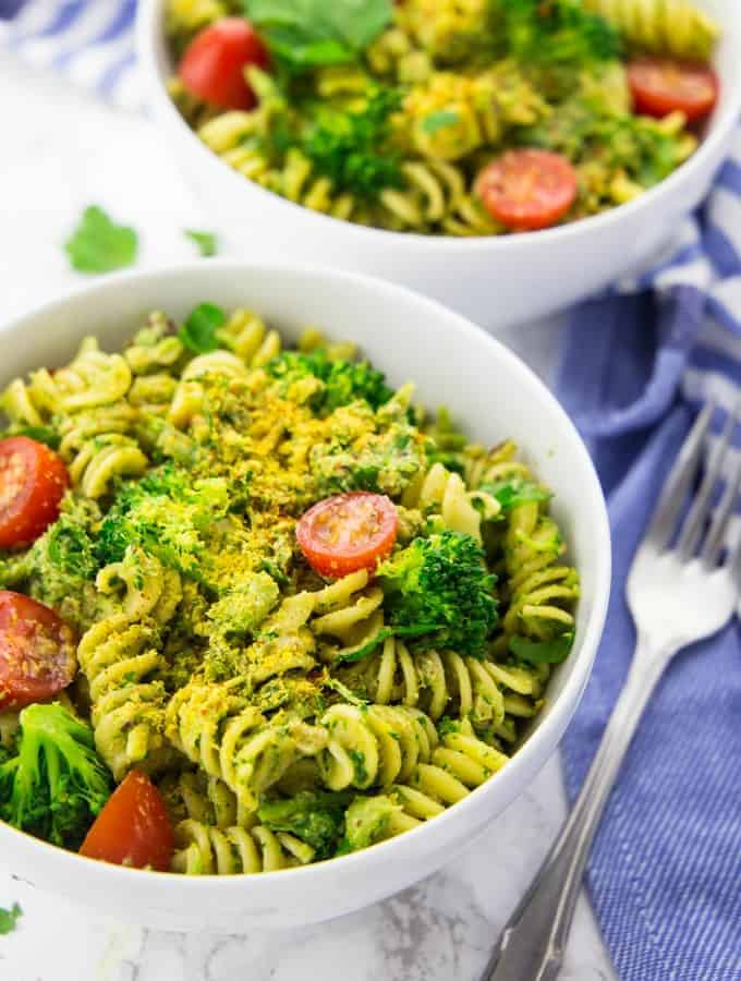 two bowls of broccoli pesto pasta with cherry tomatoes on a marble counter top with a fork and a blue napkin on the side