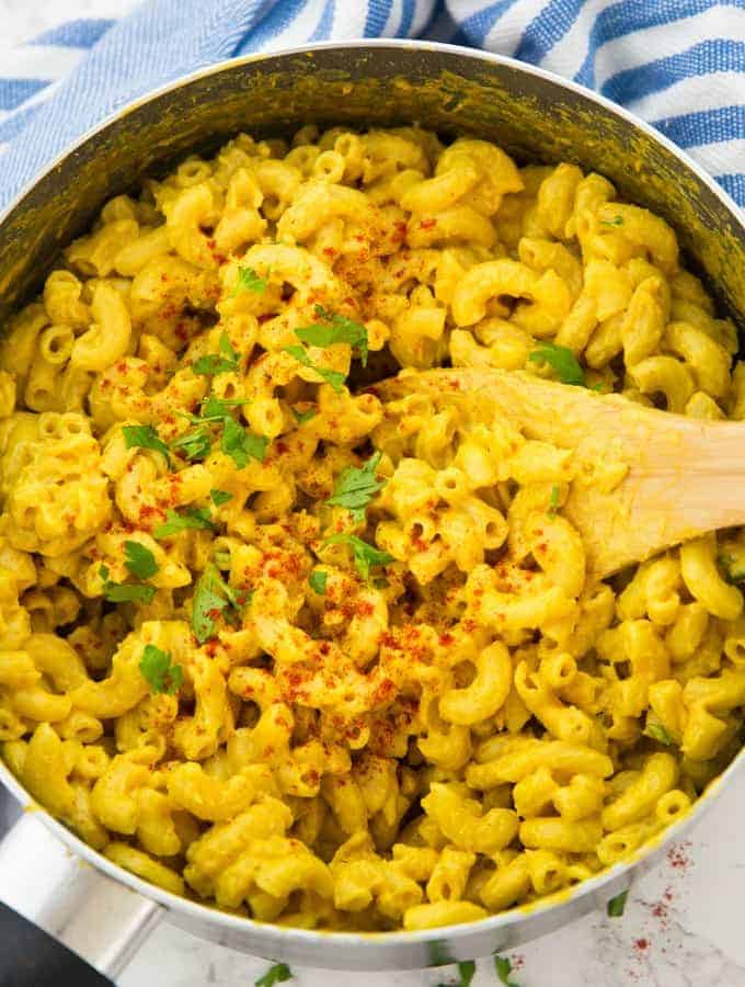 A pot of vegan mac and cheese with a wooden spoon and paprika powder and chopped parsley on top