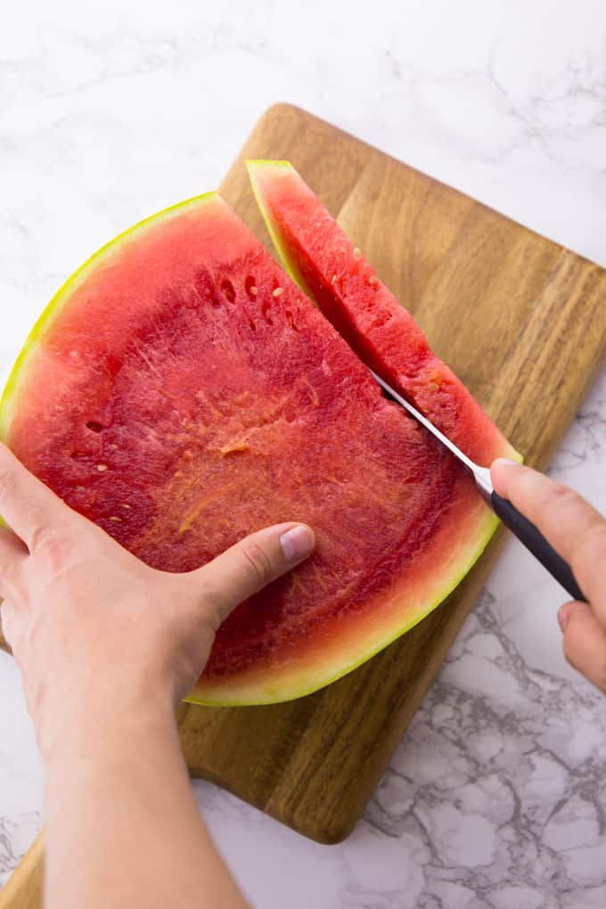 A watermelon being sliced with a large knife on a wooden board on top of a marble counter top