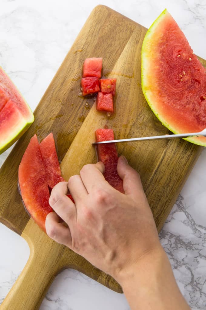 A watermelon being cut into cubes with a large knife on a wooden board on top of a marble counter top