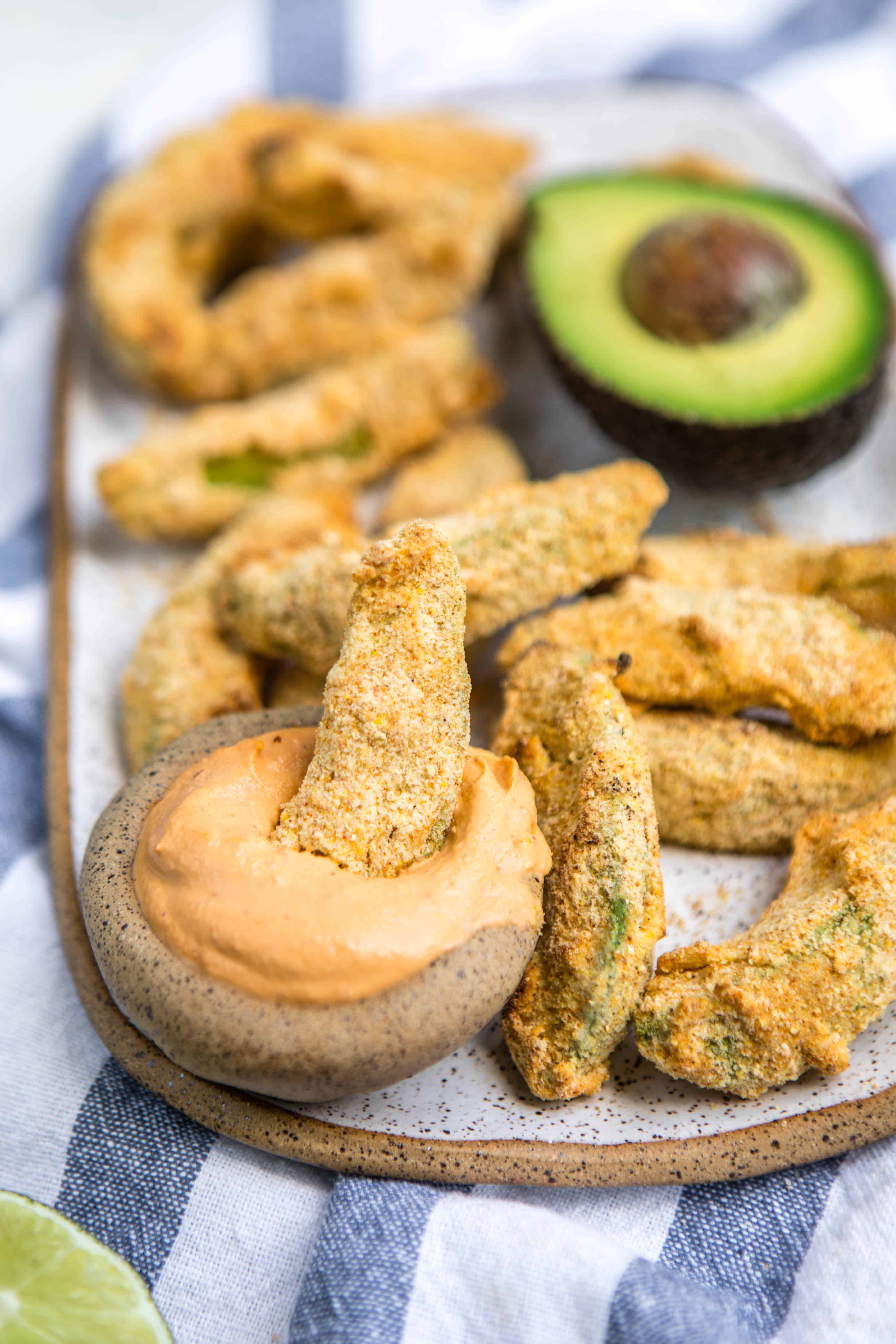 avocado fries on a grey plate on a blue and white dish cloth with an avocado half in the background a small bowl of dipping sauce in the front