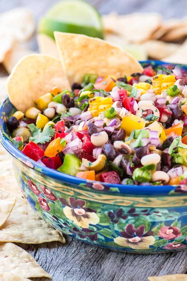 a bowl of cowboy caviar on a wooden counter top with nachos on the side and lime halves in the background