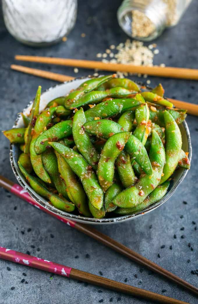 a bowl of edamame on a grey counter top with cop sticks and a bowl of salt on the side 