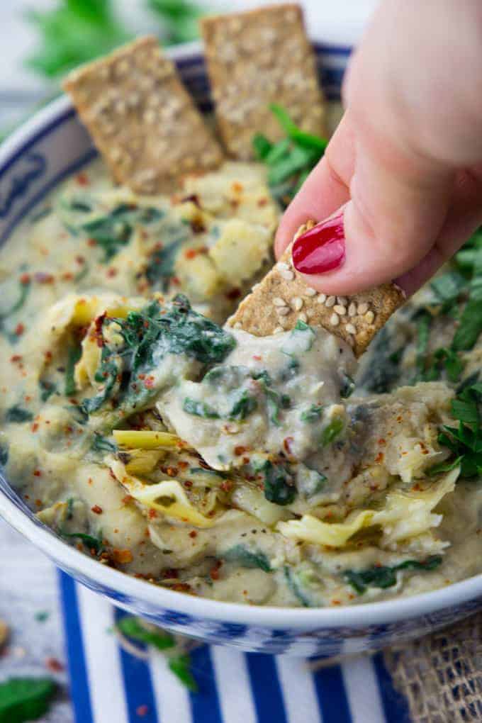 a hand dipping a sesame cracker into a bowl of vegan spinach artichoke dip on a blue napkin