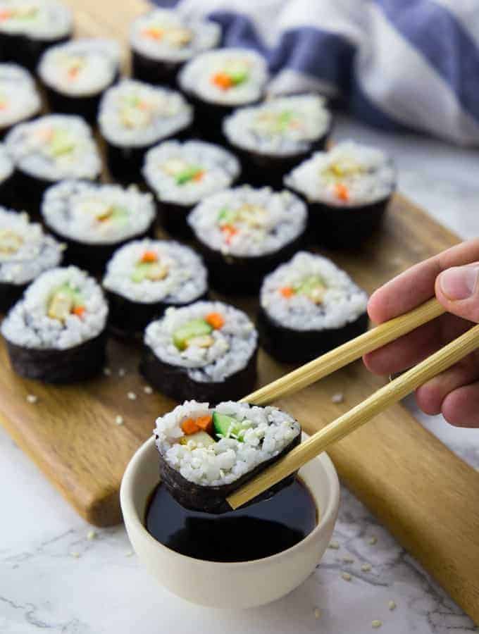 vegan sushi on a wooden board with one piece of sushi being dipped into a small bowl of soy sauce in the front