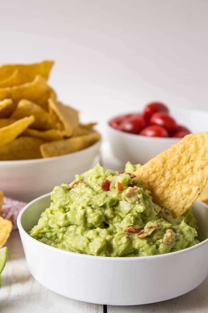 a bowl of guacamole on a white counter tip with another bowl of nachos and cherry tomatoes in the background