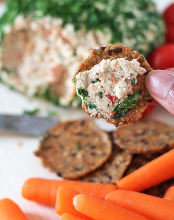 A Vegan Vegetable Almond Cheese Ball with a hand holding a cracker with cheese on it and a few snack carrots in the front
