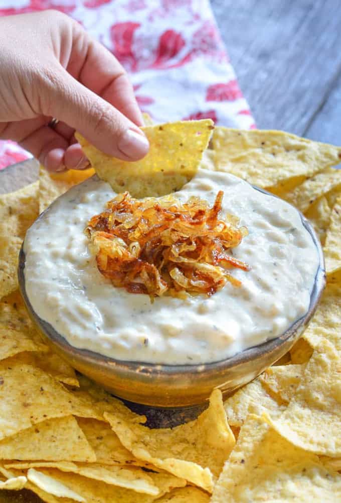 a hand dipping a nacho in a bowl of vegan garlic onion dip with more nachos around the bowl and a red table cloth in the background
