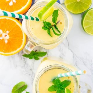 two glasses with mango smoothie on a marble counter top with orange and lime halves and mint leaves on the side