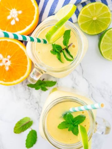 two glasses with mango smoothie on a marble counter top with orange and lime halves and mint leaves on the side