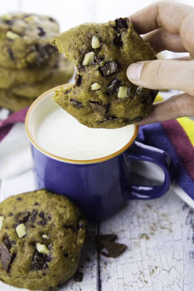 a hand holding a vegan pumpkin cookie over a blue mug with almond milk with more cookies in the background