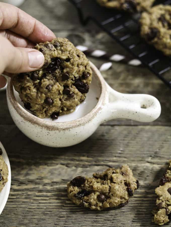 a hand dipping an oatmeal raisin cookie into a small bowl of milk with more cookies on the side