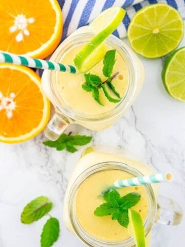 two glasses with mango smoothie on a marble counter top with orange and lime halves and mint leaves on the side
