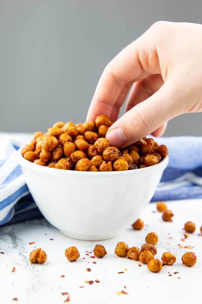 a hand eating roasted chickpeas out of a small white bowl on a marble counter top with a grey background 
