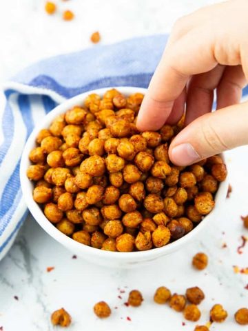 a hand eating roasted chickpeas out of a small white bowl on a marble counter top