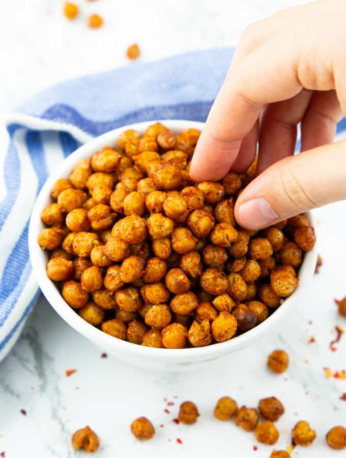 a hand eating roasted chickpeas out of a small white bowl on a marble counter top